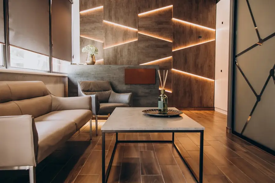 Sala de espera pequena e aconchegante, com sofá bege, mesa de centro e prateleiras de madeira com iluminação embutida